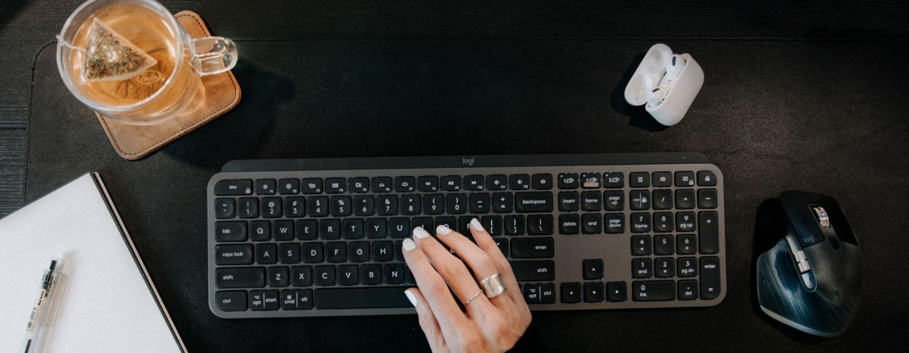 woman's hand on a keyboard surrounded by office items and a cup of tea on a coaster